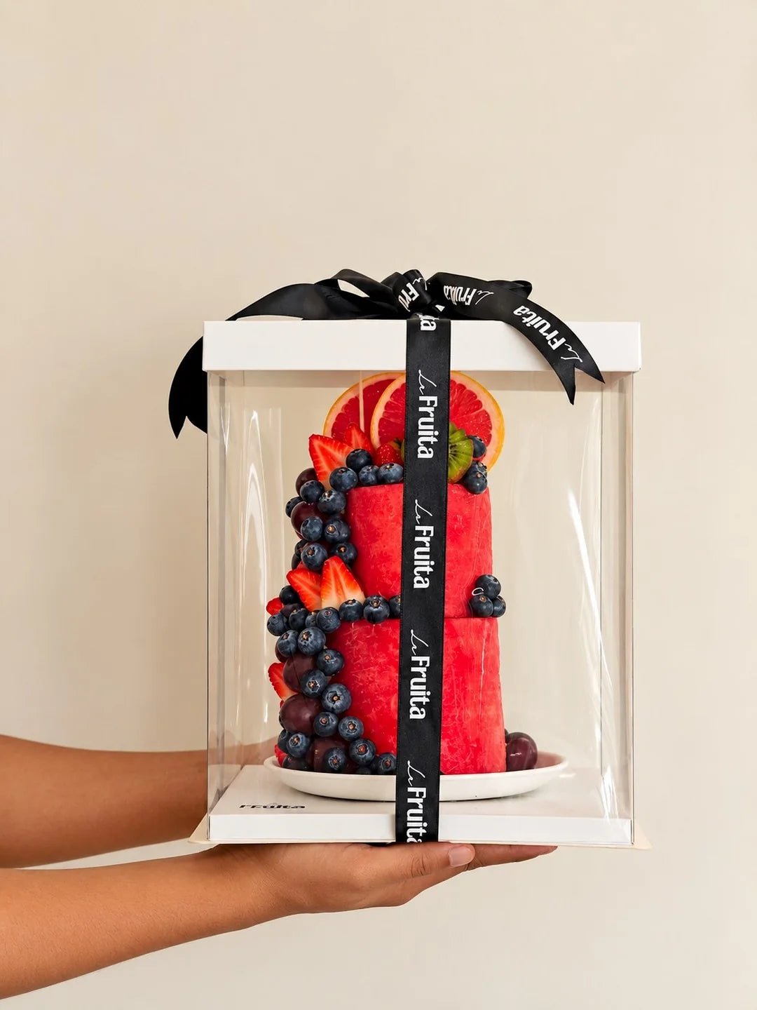 A La Fruita fruit cake in a clear presentation box with a black branded ribbon, held in hands